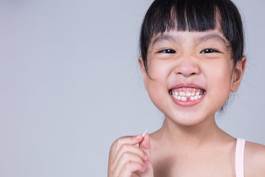 Young girl with black bangs is smiling while missing one of her front teeth.